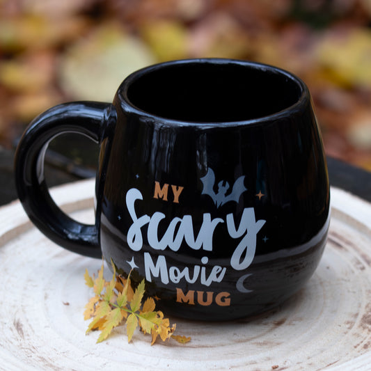 Eine schwarze Tasse mit der Aufschrift ‘My Scary Movie Mug’, verziert mit Illustrationen einer Fledermaus, Sternen und einem Mond. Die Tasse steht auf einer runden Holzscheibe, dekoriert mit gelben Herbstblättern. Der Hintergrund zeigt unscharfes herbstliches Laub.