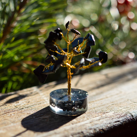 Dekoratives Bäumchen aus schwarzen Achat-Steinen mit goldfarbenem Draht auf einer sonnigen Holzoberfläche, umgeben von unscharfem, grünem Hintergrund.