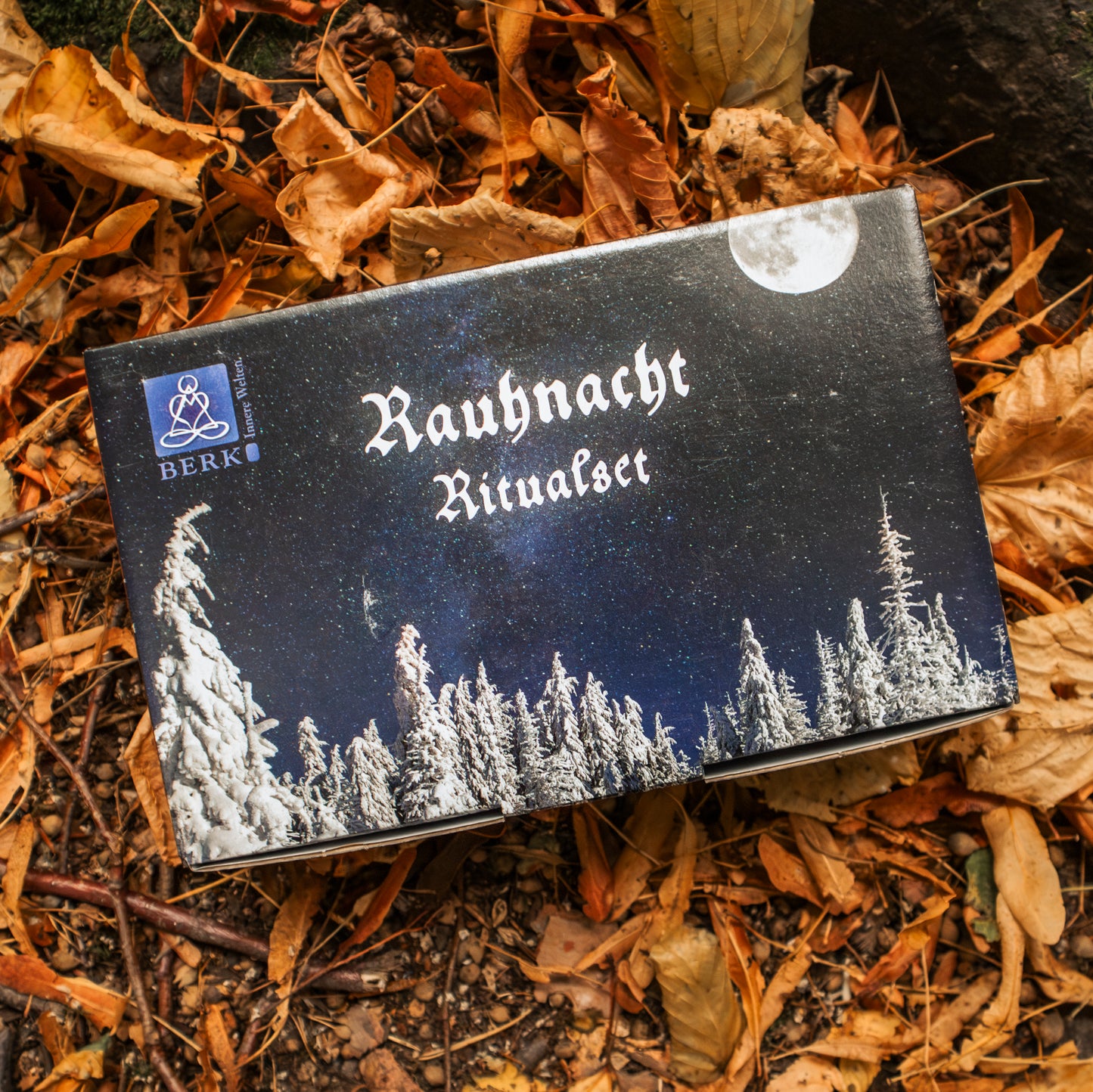 Ein Rauhnacht-Ritualset mit winterlichem Waldmotiv und Vollmond auf der Verpackung, arrangiert auf herbstlichem Blätterboden.