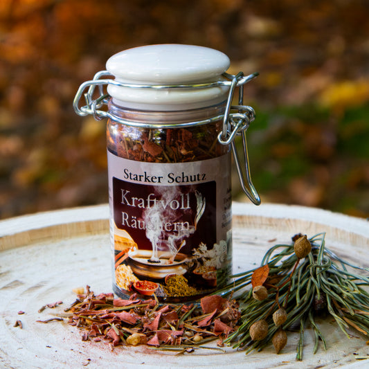 Glasbehälter mit der Aufschrift ‘Starker Schutz – Kraftvoll Räuchern’, gefüllt mit Räuchermischung, auf einer Holzscheibe arrangiert, dekoriert mit getrockneten Kräutern und losem Räucherwerk vor herbstlichem Hintergrund.