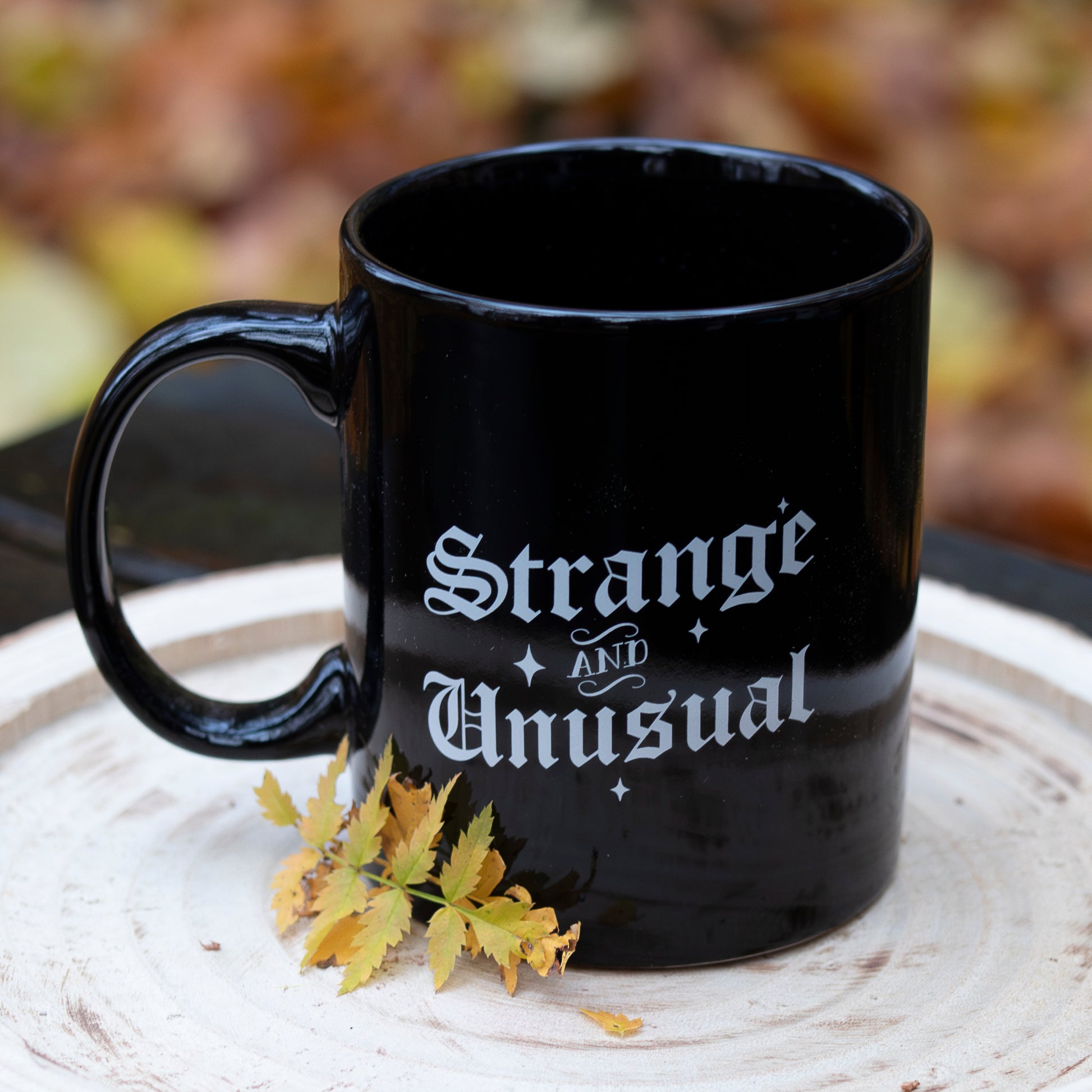 Eine schwarze Tasse mit der weißen Aufschrift ‘Strange and Unusual’ in stilisierter Schrift. Die Tasse steht auf einer runden Holzscheibe, dekoriert mit gelben Herbstblättern. Im Hintergrund ist unscharfes herbstliches Laub zu sehen.