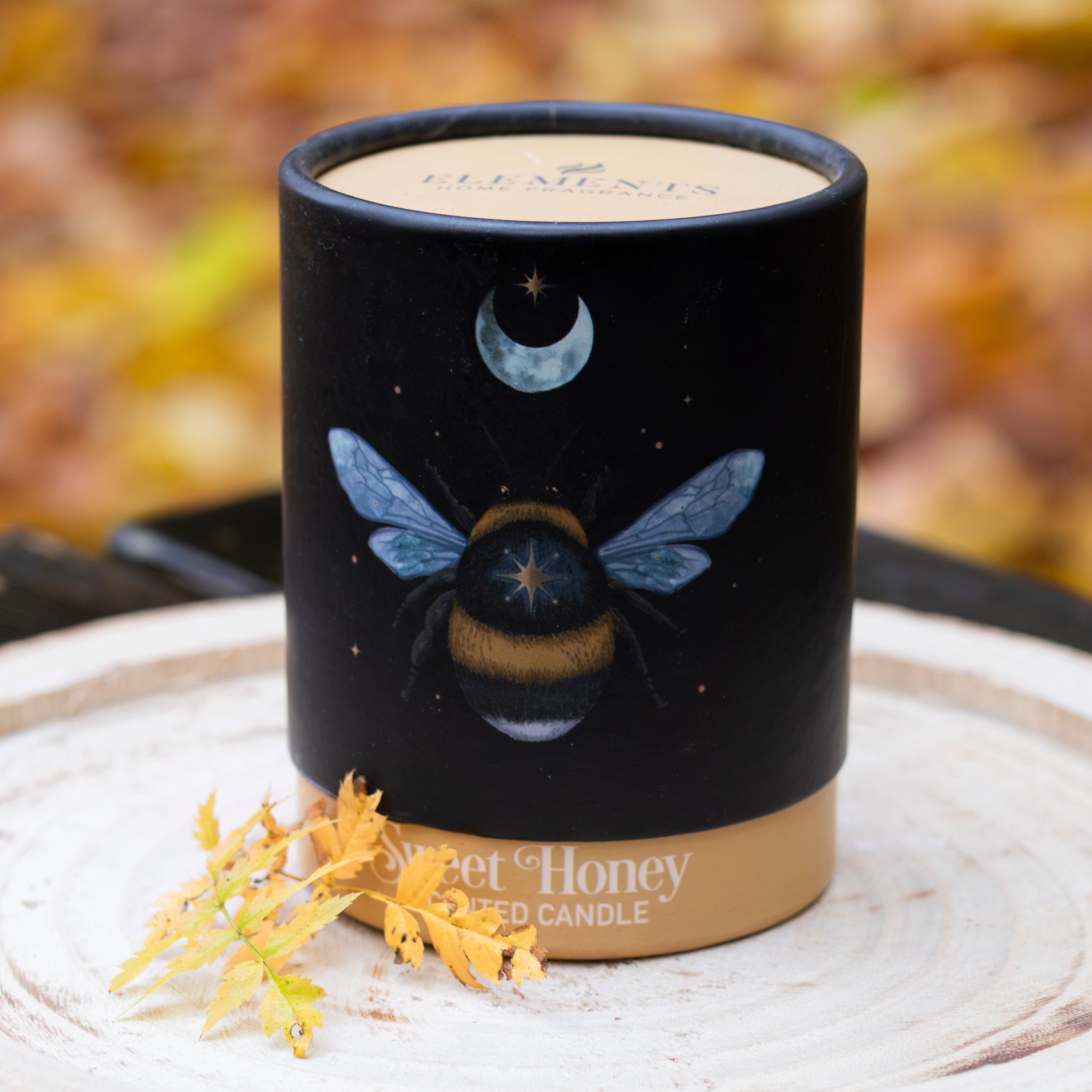 Schwarze Duftkerze mit der Aufschrift ‘Sweet Honey Scented Candle’, verziert mit einem Bienendesign und Mondsymbolen, dekoriert mit Herbstblättern auf einem rustikalen Holztablett.