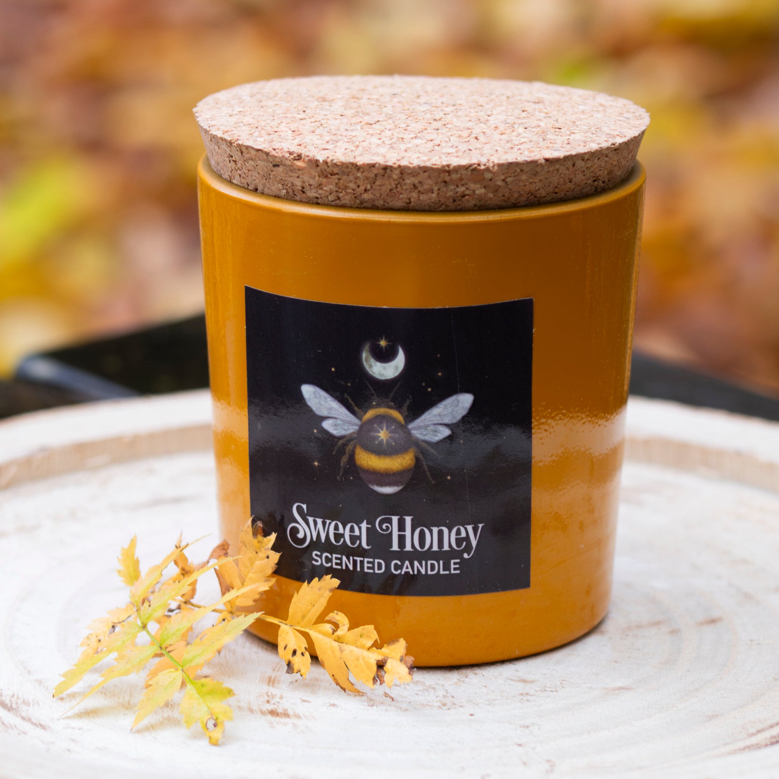 Gelbe Duftkerze mit der Aufschrift ‘Sweet Honey Scented Candle’, einem Bienendesign und einem Korkdeckel, dekoriert mit Herbstblättern auf einem rustikalen Holztablett.