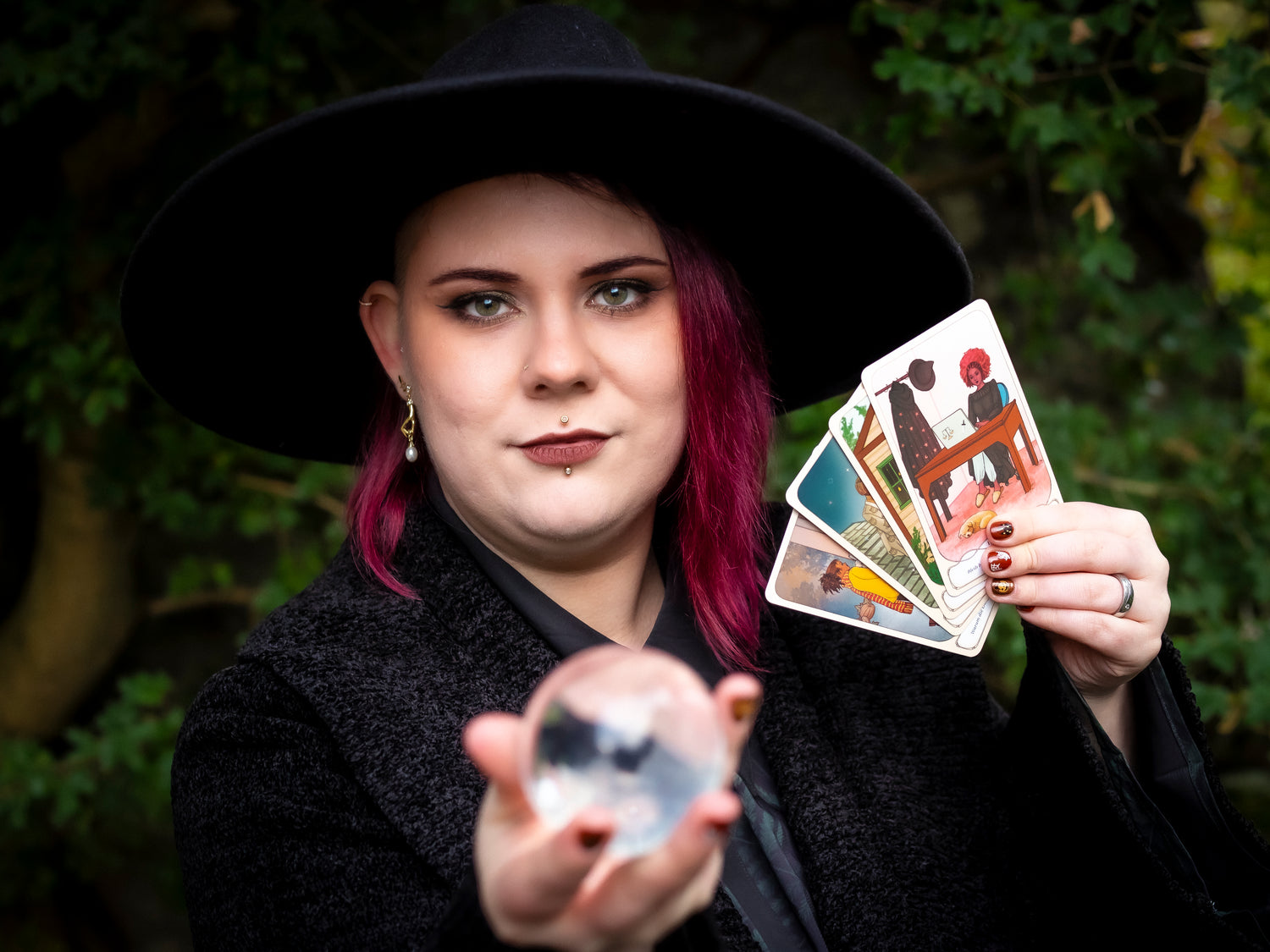 Eine Frau mit violettem Haar und schwarzem Hut hält eine Kristallkugel in der einen Hand und mehrere Tarotkarten in der anderen, vor einem Hintergrund aus grünem Laub, in einer mystischen Atmosphäre.