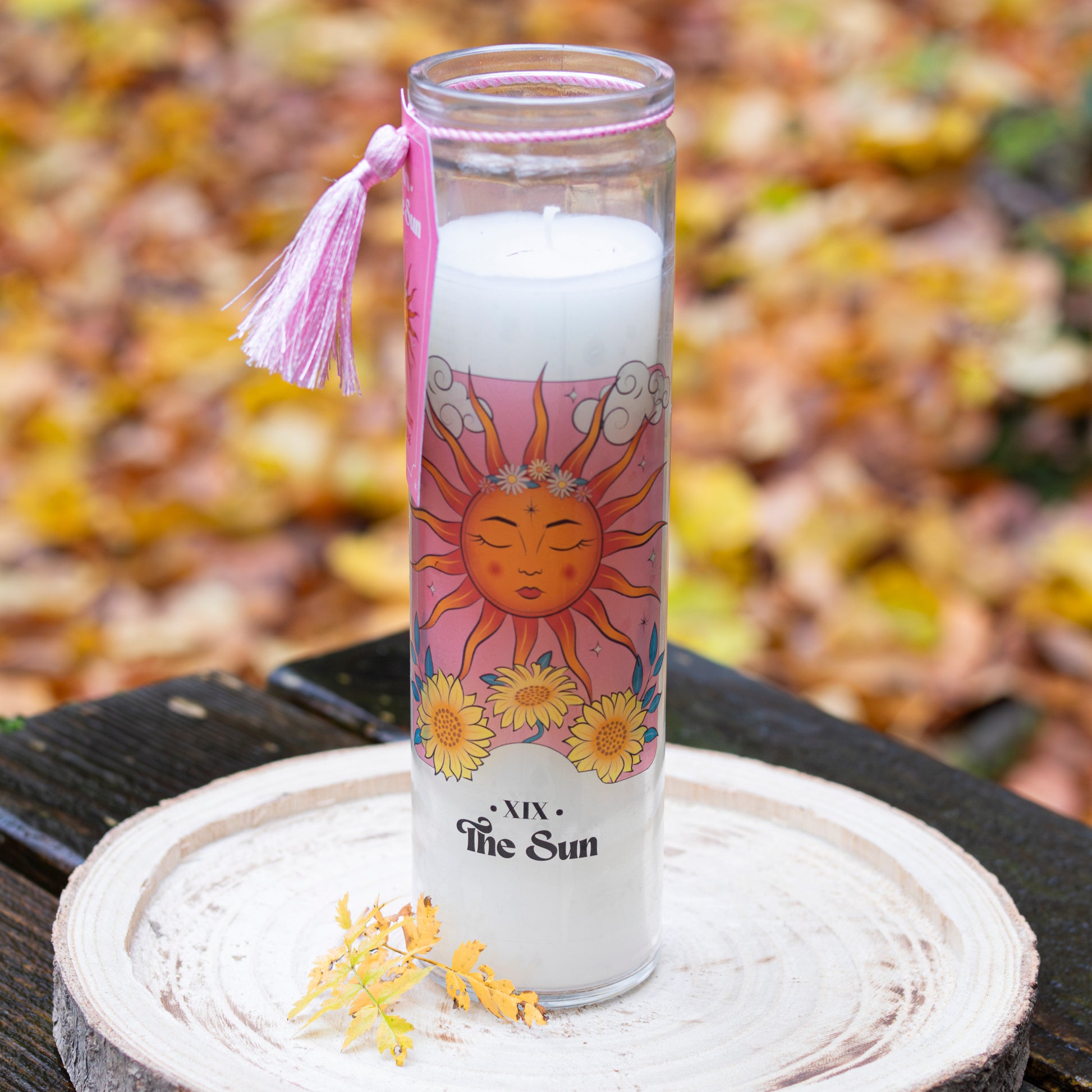 Eine weiße Kerze in einem hohen Glas mit der Aufschrift ‘The Sun’ und einer Tarotkarten-Illustration der Sonne, umgeben von Sonnenblumen und Wolken. Das Glas ist mit einer rosa Quaste verziert. Die Kerze steht auf einer runden Holzscheibe, im Hintergrund herbstliche Blätter.