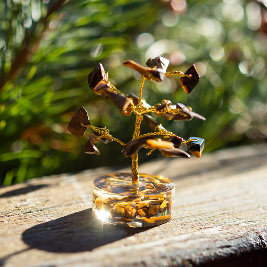 Dekoratives Bäumchen aus Tigerauge-Steinen mit goldfarbenem Draht auf einer sonnigen Holzoberfläche, umgeben von unscharfem, grünem Hintergrund.