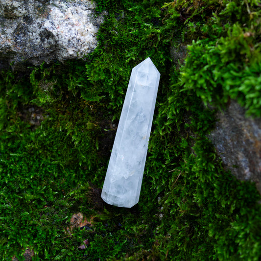 Ein klarer Bergkristall-Obelisk mit facettierten Kanten liegt auf moosbewachsenem Boden und einem Stein in natürlicher Umgebung.