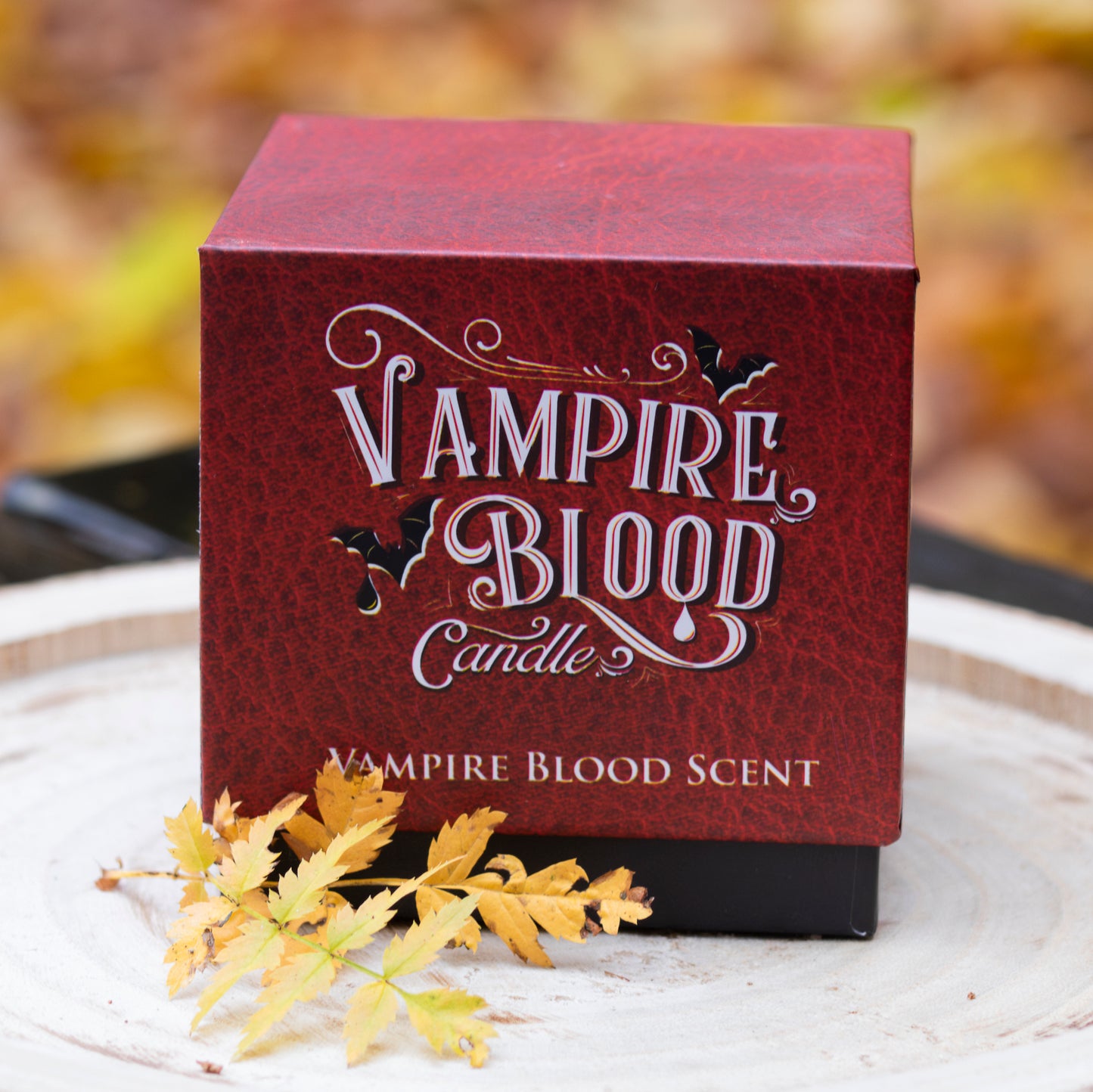 Eine elegante rote Verpackung mit der Aufschrift ‘Vampire Blood Candle’ und dekorativen Details wie Fledermäusen und verschnörkelten Schriftzügen. Die mystische und ansprechende Gestaltung deutet auf einen einzigartigen und dunklen Duft hin. Die Verpackung liegt auf einer hellen Holzscheibe, umgeben von herbstlichen Blättern, was eine saisonale Atmosphäre erzeugt.