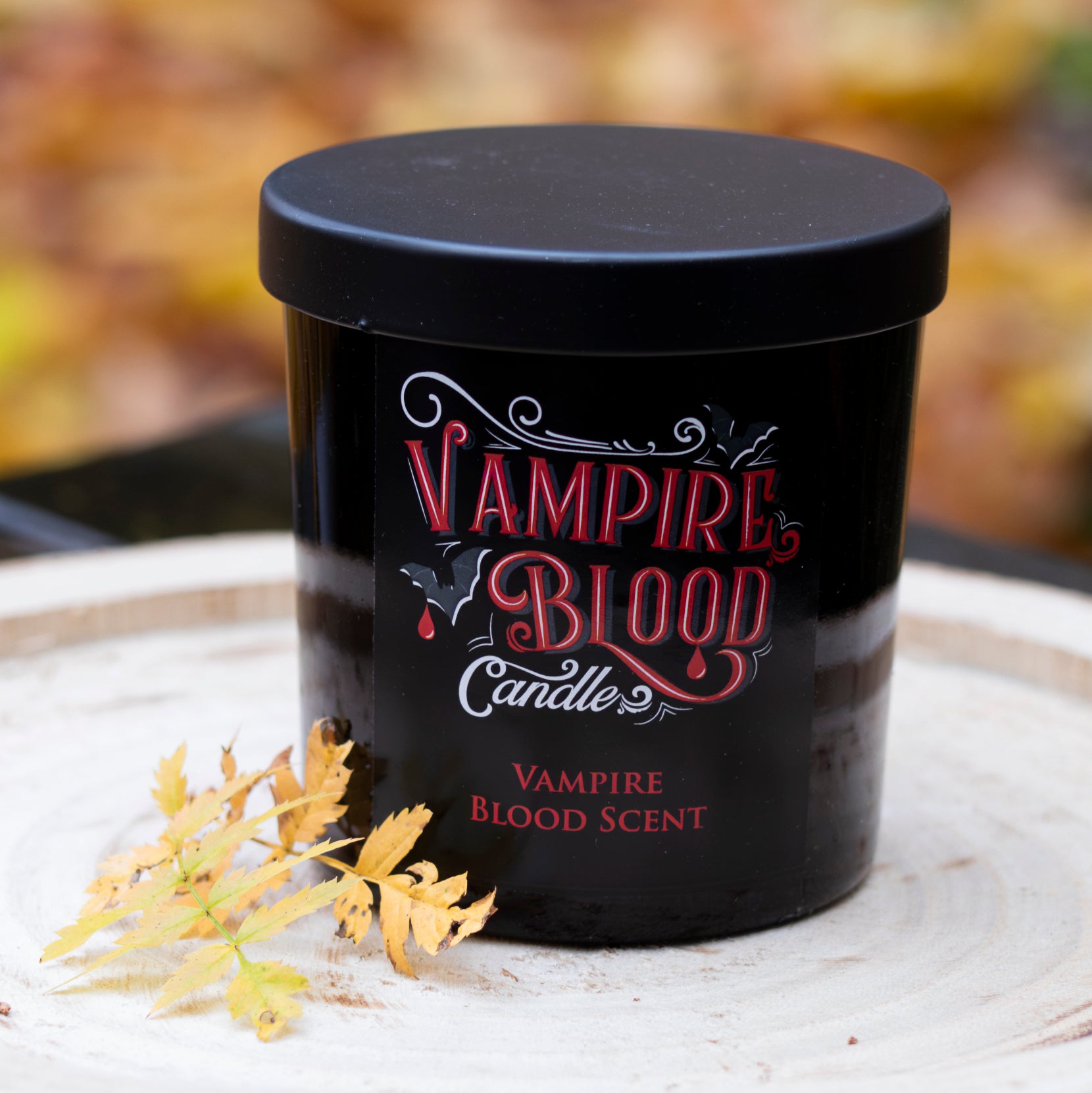 Duftkerze ‚Vampire Blood‘ in einem schwarzen Behälter mit rotem Schriftzug, dekorativ auf einer Holzscheibe platziert und mit Herbstblättern verziert.