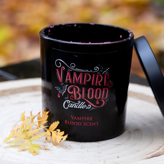 Schwarze Duftkerze mit der Aufschrift ‘Vampire Blood Candle’ in roten Buchstaben und einem schwarzen Deckel, dekoriert mit Herbstblättern auf einem rustikalen Holztablett.