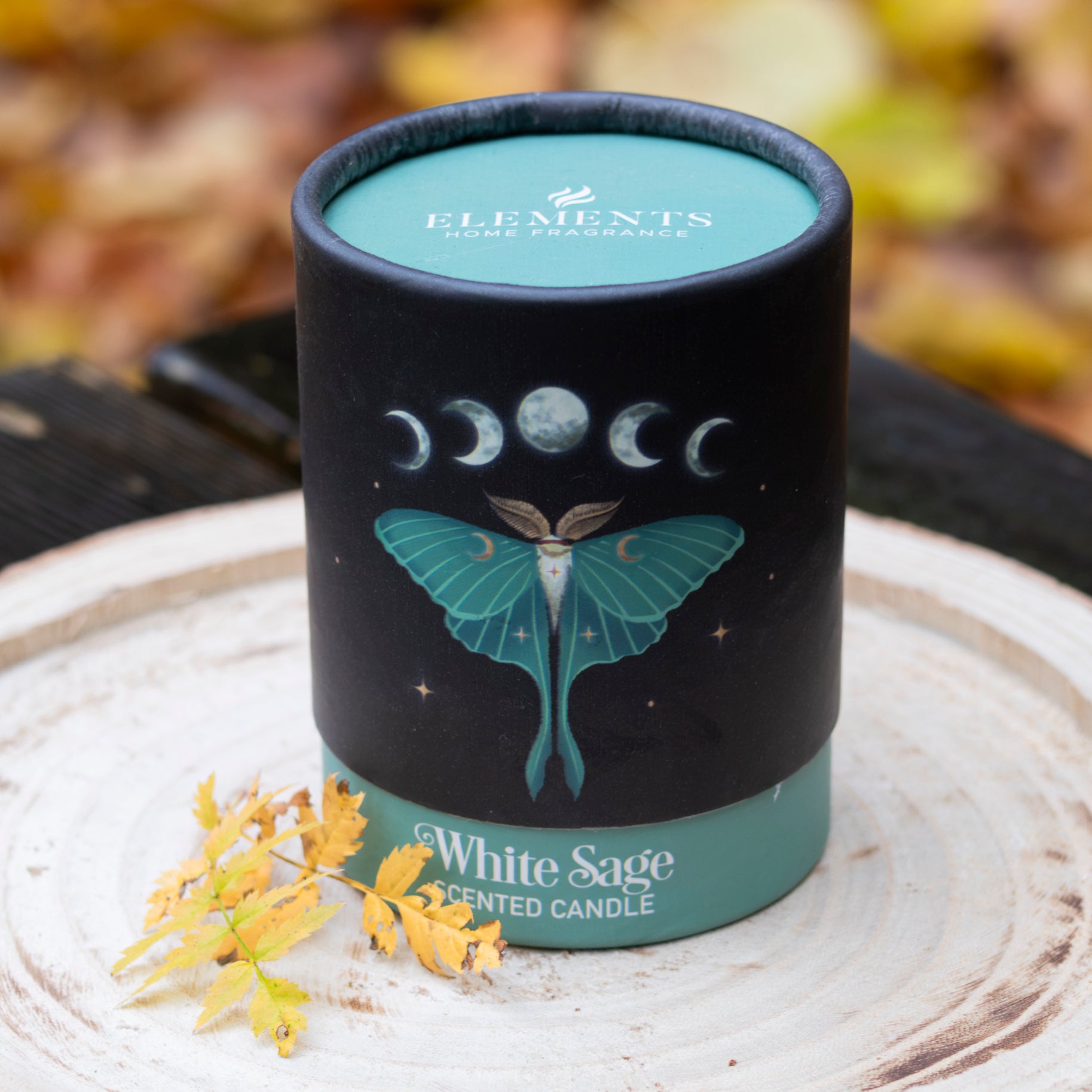 Eine elegante Duftkerze mit dem Namen ‘White Sage’ in einer hochwertigen, dunklen Verpackung. Das Design zeigt einen grünen Nachtfalter vor einer Darstellung der Mondphasen, begleitet von einer mystischen Atmosphäre. Der Behälter steht auf einer rustikalen Holzscheibe, dekoriert mit herbstlichen Blättern, und vermittelt ein Gefühl von Reinheit und natürlicher Harmonie.