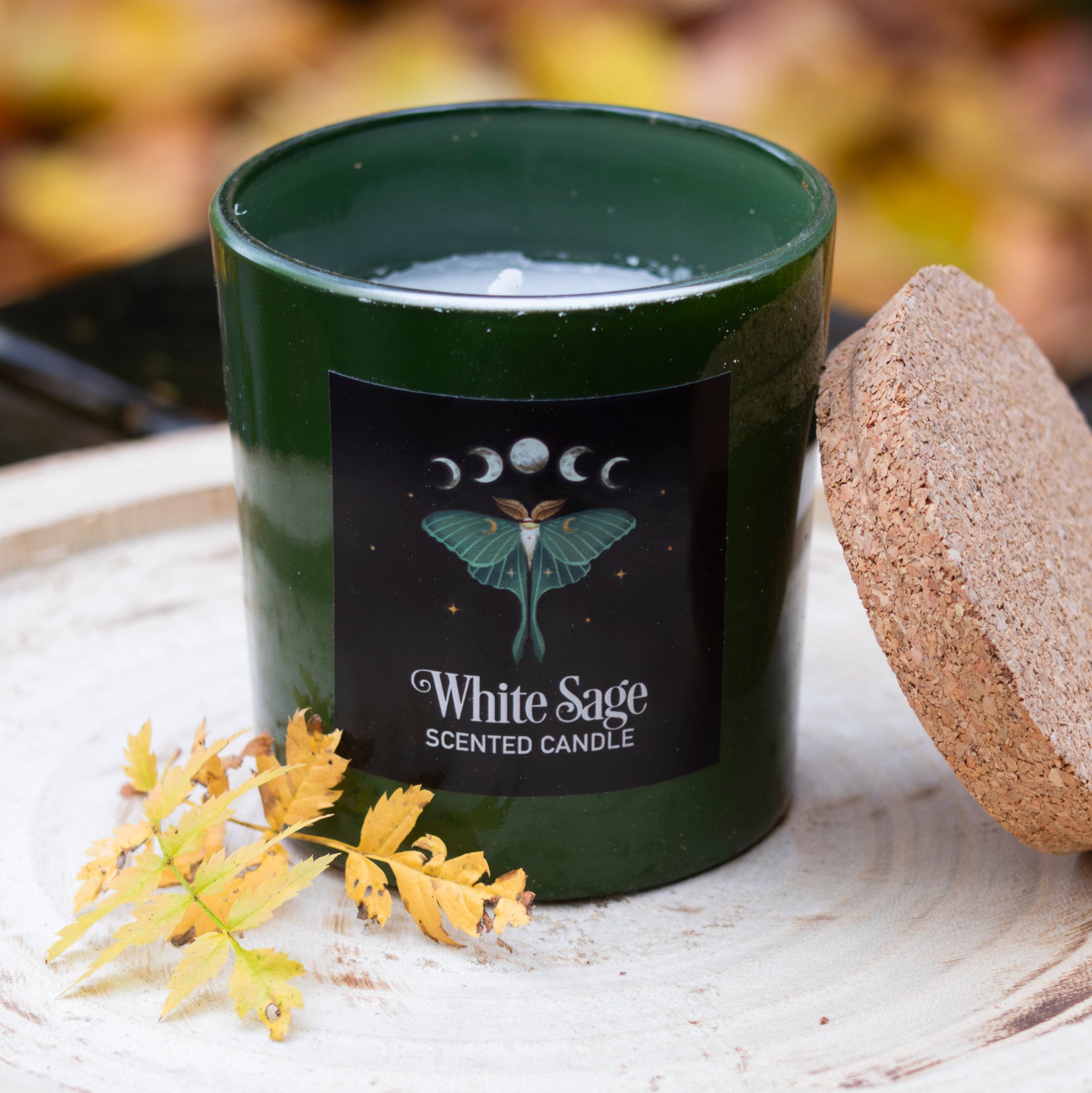 Grüne Duftkerze im Glas mit der Aufschrift ‘White Sage Scented Candle’ und einem Motten-Design, neben einem Korkdeckel und dekorativen Herbstblättern auf einem rustikalen Holztablett.