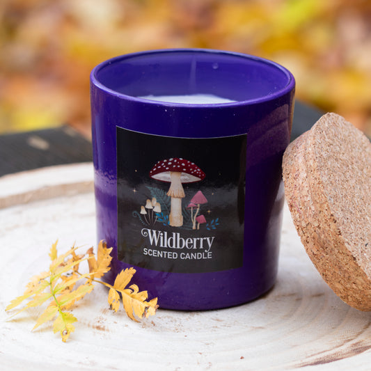 Eine tiefviolette Duftkerze in einem eleganten Glasbehälter mit einem naturbelassenen Korkdeckel. Das Etikett zeigt ein stimmungsvolles Design mit Waldpilzen und Pflanzen sowie dem Schriftzug ‘Wildberry Scented Candle’. Die Kerze steht auf einer rustikalen Holzscheibe, dekoriert mit herbstlichen Blättern, und vermittelt eine warme, naturverbundene Atmosphäre.