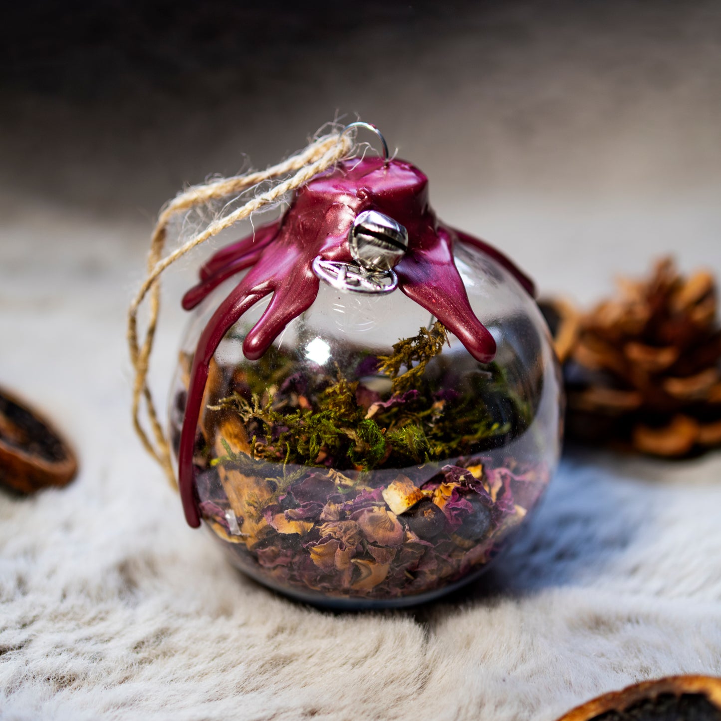 Glaskugel, gefüllt mit getrockneten Blumen, Kräutern und einer Zimtstange, dekoriert mit rotem Wachssiegel und naturfarbenem Faden. Hintergrund mit Kiefernzapfen und weicher Textur.