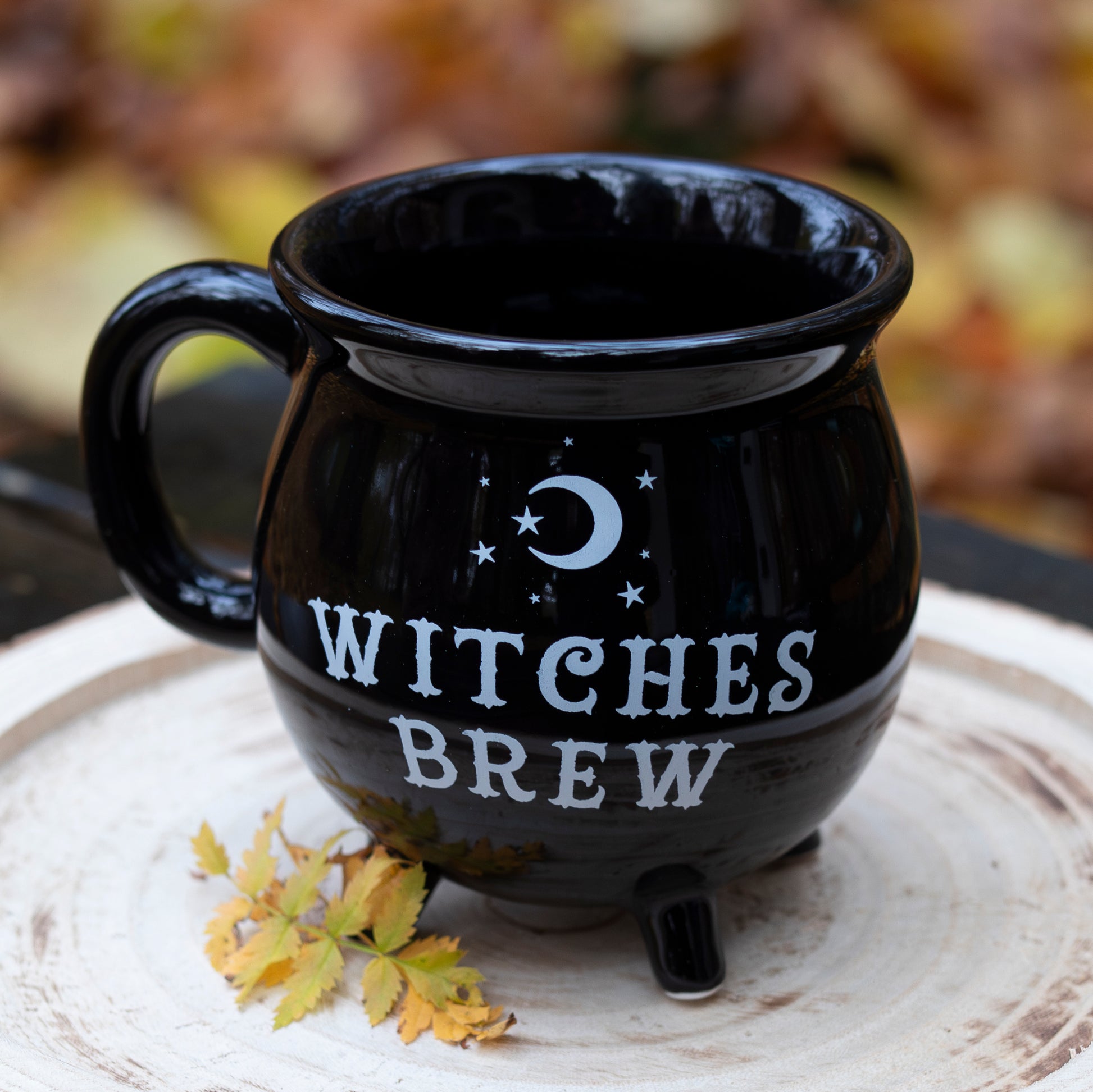 Eine schwarze Tasse in Form eines Kessels mit der weißen Aufschrift ‘Witches Brew’, verziert mit einem Mond und Sternen. Die Tasse steht auf einer runden Holzscheibe, dekoriert mit gelben Herbstblättern. Im Hintergrund ist unscharfes herbstliches Laub zu sehen.